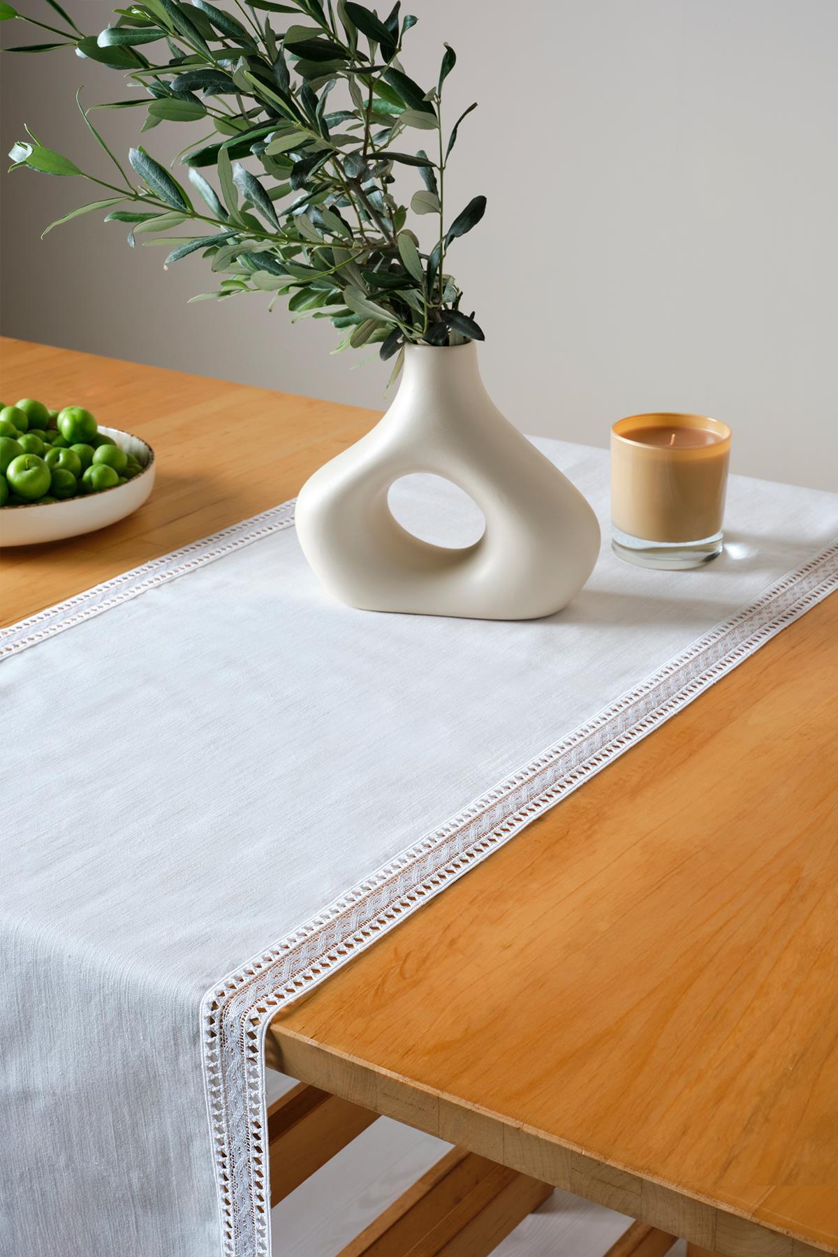 Runner da tavola in pizzo bianco su un tavolo rettangolare in legno, con vaso moderno, frutta verde e decorazione con candele.
