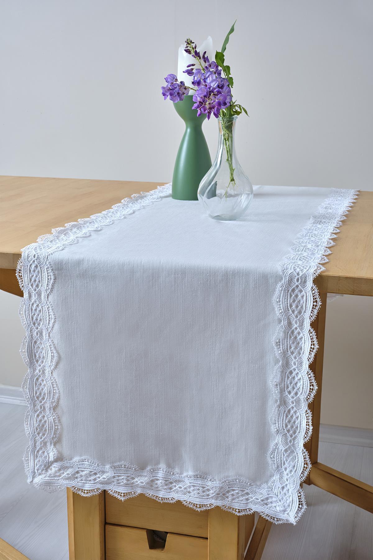 Runner bianco con bordo in pizzo, decorato con un vaso di vetro e fiori viola su un tavolo di legno