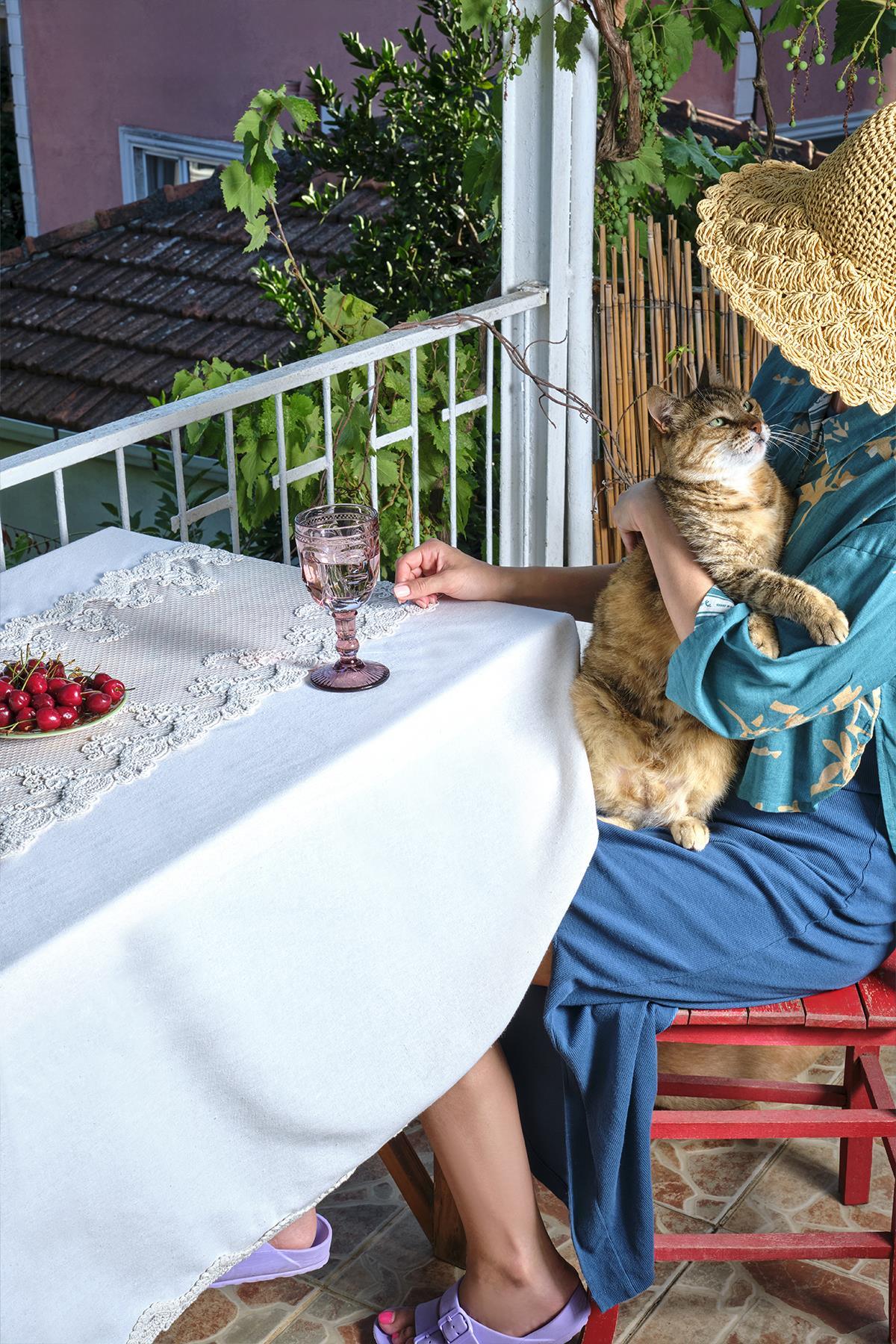 Tovaglia con ricami in pizzo su un tavolo in balcone, con sopra ciliegie e un bicchiere rosa, accanto a un gatto